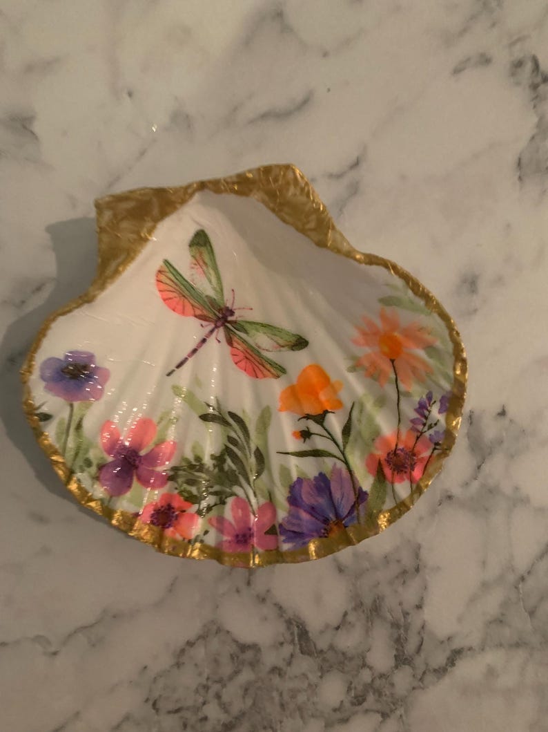 K&ouml;nnte beinhalten: Handbemalte Muschelschale mit goldfarbenem Rand. Das Innere zeigt eine Libelle und bunte Blumen in Rosa-, Lila- und Oranget&ouml;nen. Die Schale ist wei&szlig; und liegt auf einer Marmoroberfl&auml;che.