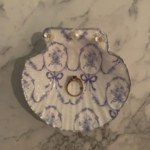 K&ouml;nnte beinhalten: Eine dekorative Muschelschalen-Ringschale mit blau-wei&szlig;em Blumenmuster. Die Schale hat einen silbernen Rand und ist mit kleinen floralen Verzierungen versehen. Ein goldener Ring mit einem Edelstein liegt in der Mitte.