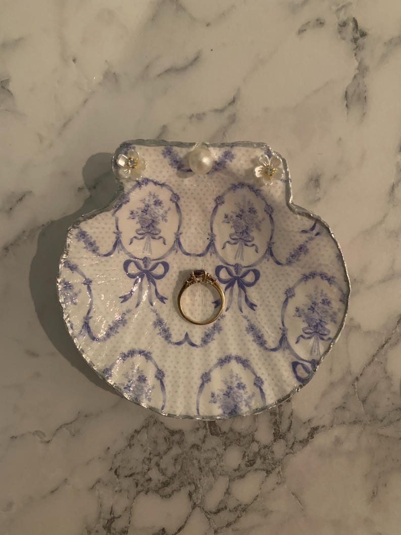 K&ouml;nnte beinhalten: Eine dekorative Muschelschalen-Ringschale mit blau-wei&szlig;em Blumenmuster. Die Schale hat einen silbernen Rand und ist mit kleinen floralen Verzierungen versehen. Ein goldener Ring mit einem Edelstein liegt in der Mitte.