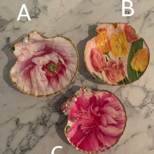 K&ouml;nnte beinhalten: Drei dekorative Muschelschalen-Tabletts. Jedes Tablett hat ein Blumendesign mit einem goldfarbenen Rand. Die Blumen sind rosa, gelb und orange. Ein Tablett hat Perlen- und Kristallverzierungen.