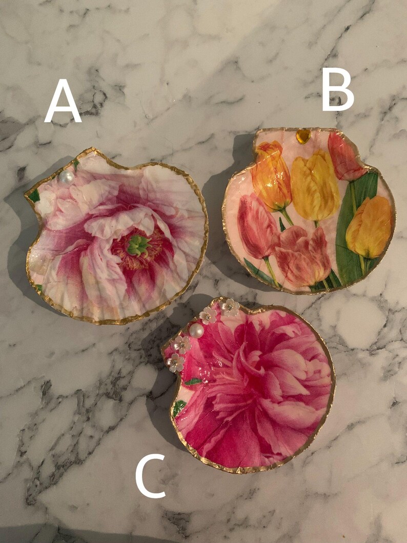 K&ouml;nnte beinhalten: Drei dekorative Muschelschalen-Tabletts. Jedes Tablett hat ein Blumendesign mit einem goldfarbenen Rand. Die Blumen sind rosa, gelb und orange. Ein Tablett hat Perlen- und Kristallverzierungen.
