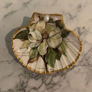 K&ouml;nnte beinhalten: Dekorative Muschelschale mit handgemalter Magnolienbl&uuml;te, gr&uuml;nen Bl&auml;ttern und Perlenakzent. Die Schale hat einen goldfarbenen Rand und einen wei&szlig;en Hintergrund mit schwacher Schrift. Ein einzigartiger Artikel f&uuml;r die K&uuml;stendekoration.
