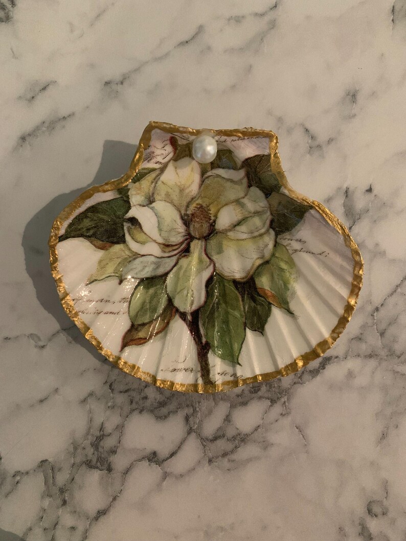 K&ouml;nnte beinhalten: Dekorative Muschelschale mit handgemalter Magnolienbl&uuml;te, gr&uuml;nen Bl&auml;ttern und Perlenakzent. Die Schale hat einen goldfarbenen Rand und einen wei&szlig;en Hintergrund mit schwacher Schrift. Ein einzigartiger Artikel f&uuml;r die K&uuml;stendekoration.