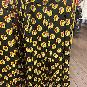 May include: Black pants with a repeating pattern of a cartoon beaver head inside a yellow circle. The beaver is wearing a red hat. The pants are hanging on a white hanger. The size XL is visible on the hanger.