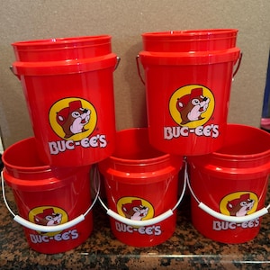 May include: Six red plastic buckets with white handles are displayed. Each bucket has a yellow and brown logo and the white text "BUC-EE'S". The buckets are stacked and arranged on a countertop.