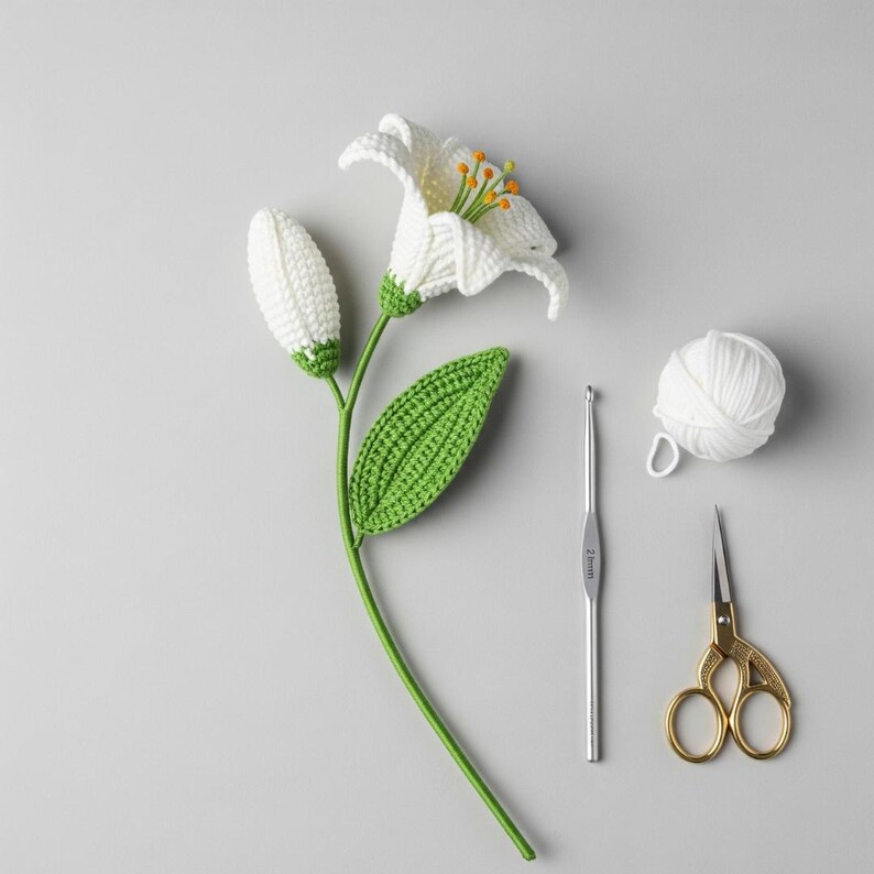 May include: A handmade crochet lily flower with a white bloom and bud, green stem and leaf, and yellow stamens. A crochet hook, a ball of white yarn, and gold scissors are also present.