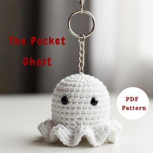 Może przedstawiać: Brelok z białym szydełkowym duchem z czarnymi guzikami i pofałdowanym dołem. Tekst "The Pocket Ghost" jest w kolorze czerwonym nad duchem. Dołączony jest srebrny łańcuszek i brelok. Słowa "PDF Pattern" znajdują się w białym okręgu.