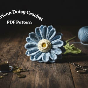 Op de afbeelding: Een gehaakte blauwe madelief met een crèmekleurig centrum en groene stengel ligt op een houten oppervlak. De tekst "African Daisy Crochet PDF Pattern" staat boven de bloem. Een bol blauw garen, een schaar en naaiattributen zijn ook zichtbaar.