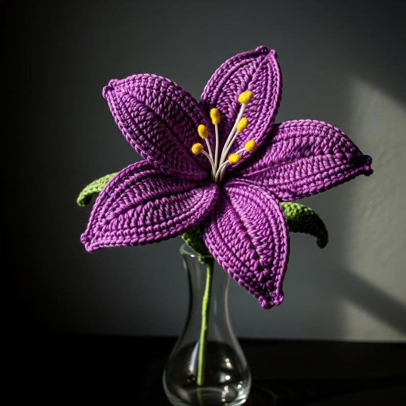May include: A handcrafted, purple crochet flower with yellow stamens and green leaves. The flower is displayed in a clear glass vase. The petals are detailed with visible crochet stitches, creating a textured appearance. The flower's stem is green.