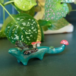 May include: A small teal ceramic creature figurine featuring dark, speckled wings, two tiny white-stemmed red mushrooms on its back, and a single red mushroom with white spots perched on the tip of its long, curved tail.