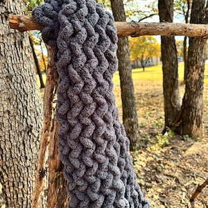 May include: A chunky, hand-knitted gray blanket is draped over a tree branch. The blanket's thick, textured weave is visible, showcasing the soft, plush yarn. The background features trees and a grassy area, suggesting an outdoor setting.