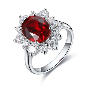 Può includere: Un anello d'argento con una grande gemma rosso scuro di taglio ovale, circondata da pietre più piccole, chiare e di taglio rotondo. La fascia in argento lucido sostiene l'incastonatura, che forma un alone floreale. Un gioiello.