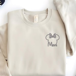 May include: A cream-coloured sweatshirt featuring a grey embroidered Minnie Mouse head and bow, with the word "Mom" beneath. The sweatshirt is laid flat, with blue jeans and a black and white striped scarf in the background.