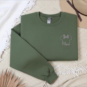 May include: An olive green sweatshirt with a crew neck and long sleeves. The design features a Minnie Mouse head outline with a bow and the word "Mom" embroidered in silver thread. The sweatshirt is folded on a cream-coloured blanket.