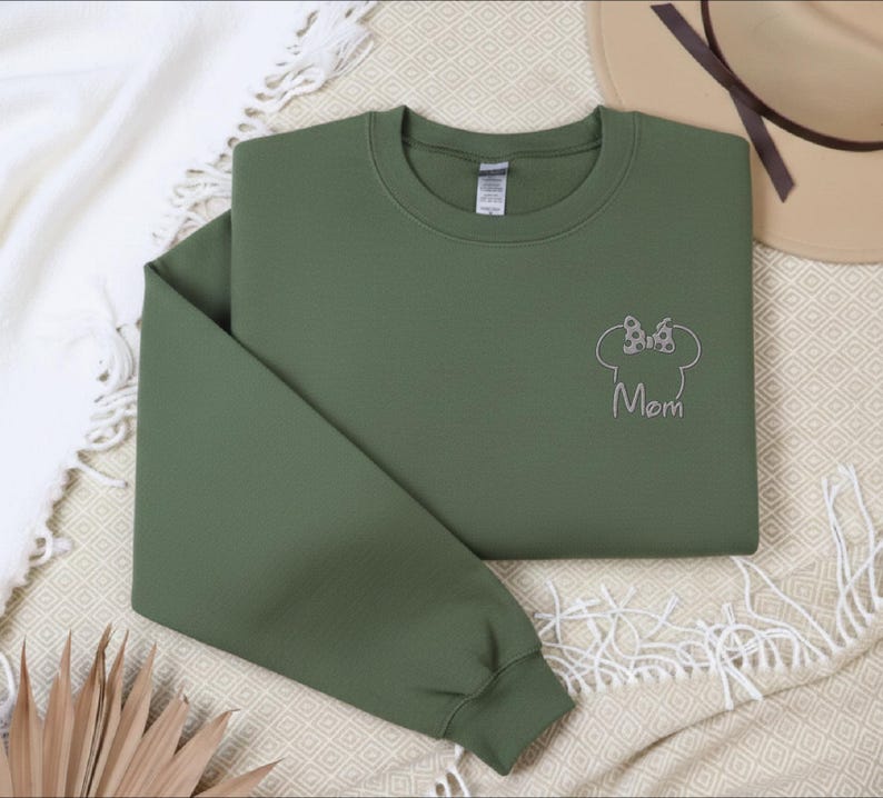 May include: An olive green sweatshirt with a crew neck and long sleeves. The design features a Minnie Mouse head outline with a bow and the word "Mom" embroidered in silver thread. The sweatshirt is folded on a cream-coloured blanket.