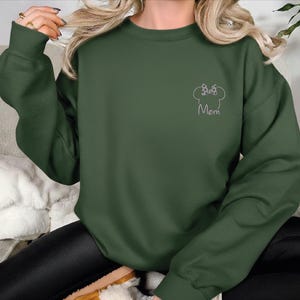 May include: Dark green sweatshirt featuring a white outline of a chef's hat and the word "Mom". The long-sleeved top is made of a soft material.