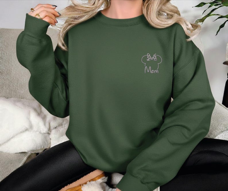 May include: Dark green sweatshirt featuring a white outline of a chef's hat and the word "Mom". The long-sleeved top is made of a soft material.