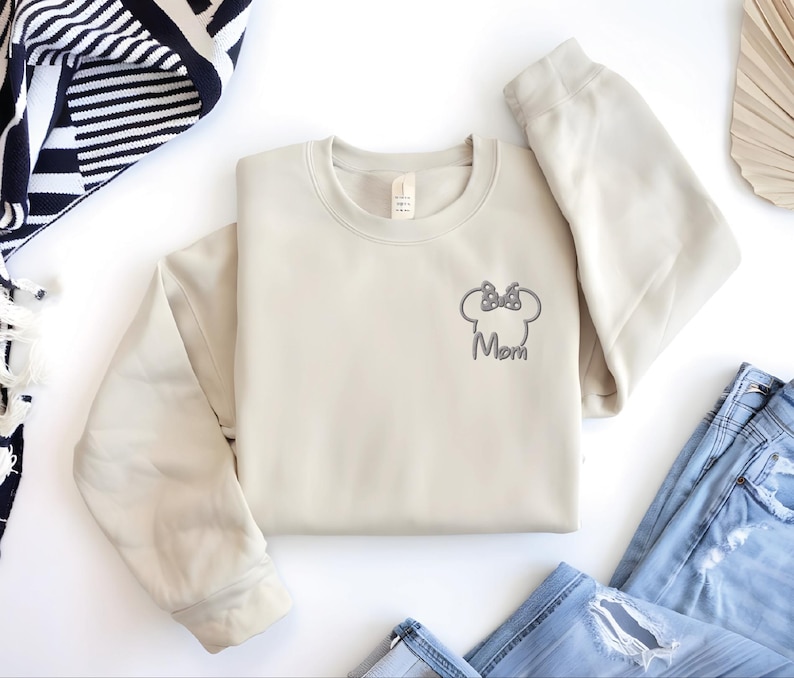 May include: A cream-coloured sweatshirt featuring a grey embroidered Minnie Mouse head and bow, with the word "Mom" beneath. The sweatshirt is laid flat, with blue jeans and a black and white striped scarf in the background.