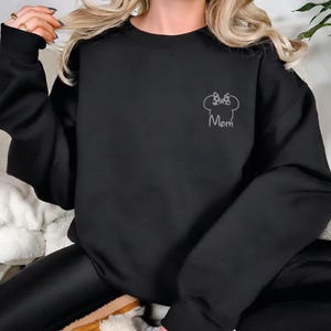 May include: Black sweatshirt featuring a white outline of a bow and mouse ears, with the word "Mom" beneath. The sweatshirt has long sleeves and a crew neck. The design is on the upper left chest area.