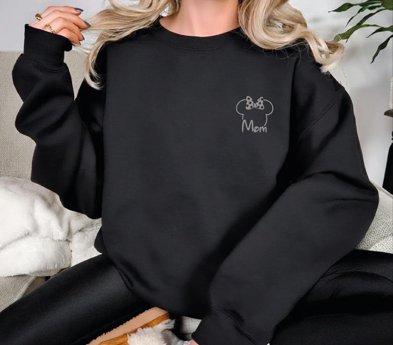 May include: Black sweatshirt featuring a white outline of a bow and mouse ears, with the word "Mom" beneath. The sweatshirt has long sleeves and a crew neck. The design is on the upper left chest area.