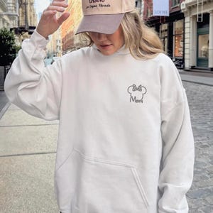 May include: A white hooded sweatshirt with a front pocket and a hood. The sweatshirt has a small embroidered design of a bow and the word "Mom". A tan baseball cap with text is worn.
