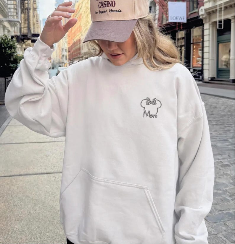 May include: A white hooded sweatshirt with a front pocket and a hood. The sweatshirt has a small embroidered design of a bow and the word "Mom". A tan baseball cap with text is worn.