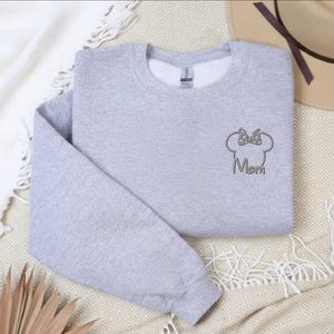 May include: A light grey sweatshirt featuring a Minnie Mouse head design and the word "Mom". The sweatshirt is folded and placed on a patterned surface, with a hat and a white blanket visible.