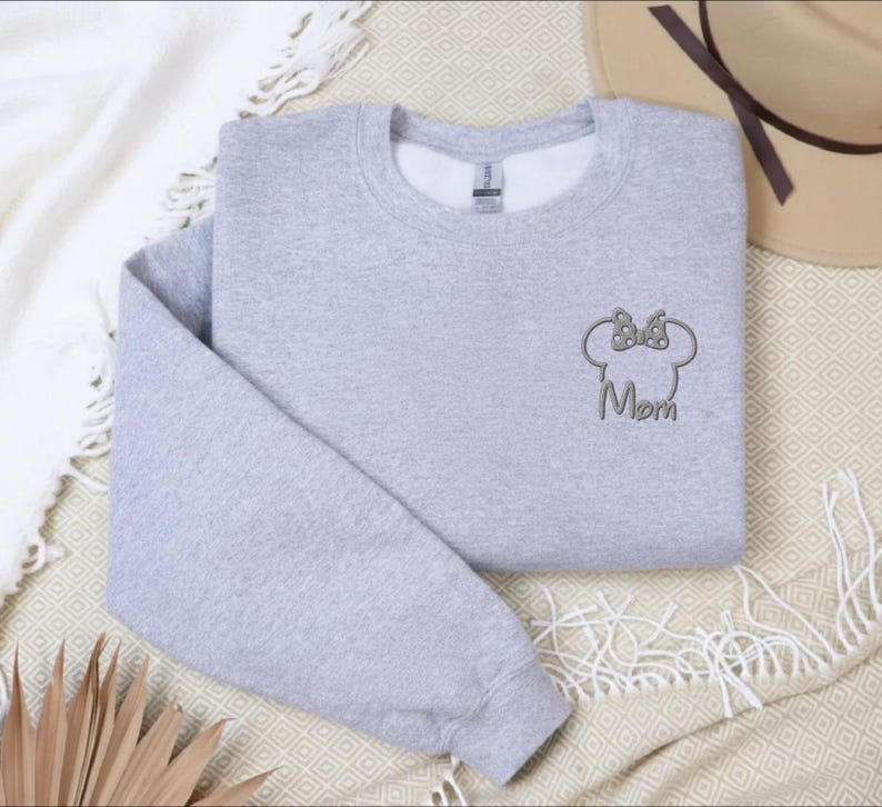 May include: A light grey sweatshirt featuring a Minnie Mouse head design and the word "Mom". The sweatshirt is folded and placed on a patterned surface, with a hat and a white blanket visible.