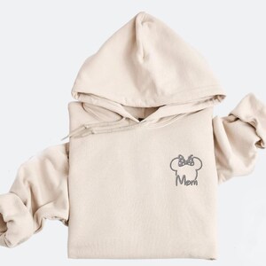May include: Beige hoodie with a drawstring hood and long sleeves. The front features a grey embroidered Minnie Mouse head with a bow and the word "Mom" underneath. The hoodie is made of a soft material.