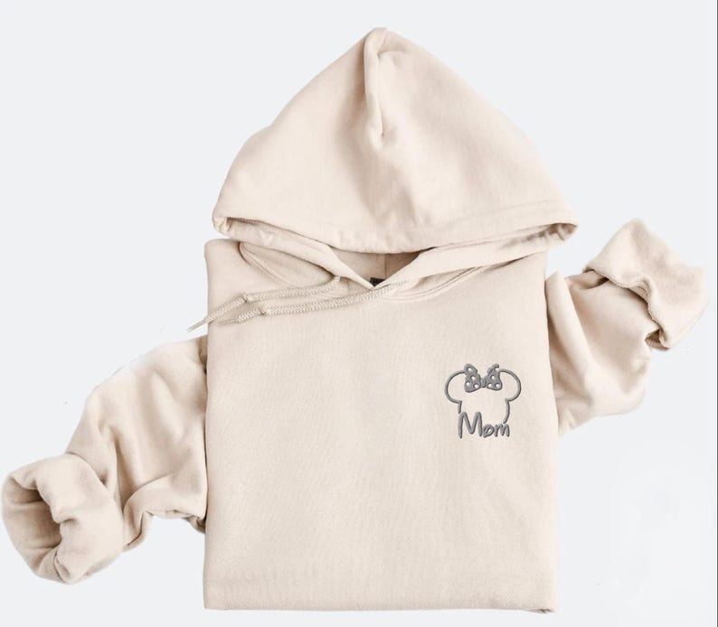 May include: Beige hoodie with a drawstring hood and long sleeves. The front features a grey embroidered Minnie Mouse head with a bow and the word "Mom" underneath. The hoodie is made of a soft material.