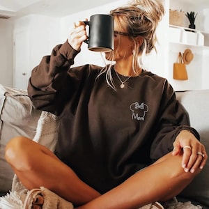 May include: A dark brown sweatshirt with a white bow and the word "Mom". A black mug is held up. The long-sleeved sweatshirt is made of a soft material. The graphic is on the front. The person is sitting down.
