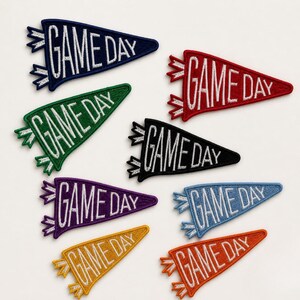 May include: A collection of eight triangular pennant-shaped embroidered patches in various colors including navy, red, green, black, purple, light blue, yellow, and orange. Each patch features the white embroidered text GAME DAY.