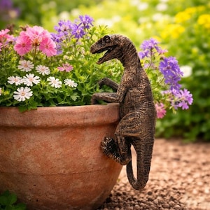 Puede incluir: Una estatua de jardín de dinosaurio de color bronce agarrada al borde de una maceta de terracota llena de flores rosas, moradas y blancas. La estatua tiene una superficie texturizada y está en posición de escalar.