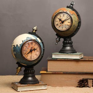 May include: Two antique globe-shaped clocks with a vintage aesthetic. The clocks feature a world map design with Roman numeral clock faces. They are set on black bases and rest on small books, with a neutral background.