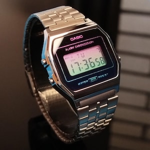 May include: A silver-toned Casio digital watch with a rectangular face and a matching metal band. The watch face displays the time, day, and date in green digits. The watch is labeled "CASIO ALARM CHRONOGRAPH" and "WATER RESIST".