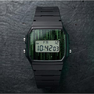 Puede incluir: Reloj digital negro con una pantalla verde estilo matriz. La esfera del reloj muestra la hora, el día y la fecha. El reloj tiene una correa y botones negros. El fondo es una superficie oscura y texturizada.