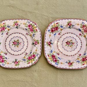 Puede incluir: Dos platos cuadrados con bordes festoneados. Cada plato presenta un estampado floral con flores rosas, azules y amarillas sobre fondo blanco. Los platos tienen un ribete dorado y un diseño floral central.