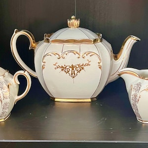 May include: A three-piece tea set in cream with gold trim. The set includes a teapot, a sugar bowl with a lid, and a creamer. The teapot has a rounded body and a gold finial. The set is decorated with gold floral patterns.