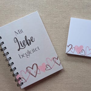 Op de afbeelding: Een wit spiraalgebonden notitieboek en een notitieblok. Het notitieboek bevat de Duitse tekst "Mit Liebe begleitet" en een rand van geschetste harten in roze en bruin. Het notitieblok heeft een vergelijkbare hartrand. Zwarte spiraalbinding.