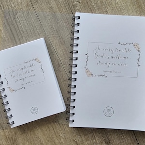 Peut inclure: Deux cahiers à spirales blancs. Le plus grand affiche le texte "In every trouble God is with me - strong an near" dans un cadre floral. Le plus petit a le même design. Les deux sont sur une surface en bois.