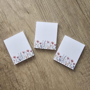 Könnte beinhalten: Drei weiße Notizblöcke mit floralem Design. Das florale Design zeigt rosa, lila und gelbe Blumen mit grünen Stielen und Blättern. Die Notizblöcke sind auf einer Holzoberfläche angeordnet.