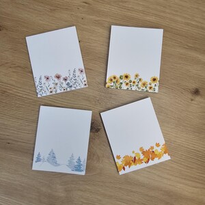 Könnte beinhalten: Vier weiße Notizblöcke mit saisonalen Designs. Einer zeigt Wildblumen, ein anderer Sonnenblumen, ein dritter Winterbäume und der letzte Herbstblätter. Die Notizblöcke sind auf einer Holzoberfläche angeordnet.