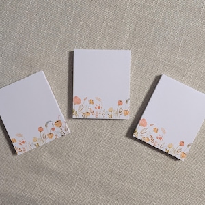 Könnte beinhalten: Drei weiße Notizblöcke mit einem Blumendesign am unteren Rand. Die Aquarellblumen sind in Pfirsich-, Gelb- und Brauntönen gehalten. Die Notizblöcke sind auf einer neutralfarbenen Stoffoberfläche angeordnet.