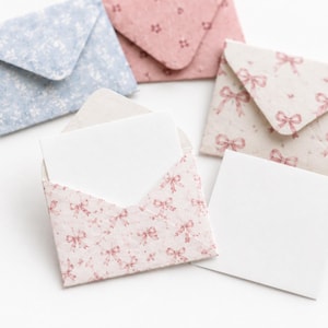 May include: A collection of small paper envelopes featuring various patterns including a light blue floral print, a solid dusty pink, and a cream envelope with a repeating pink bow design, accompanied by two blank white square note cards.