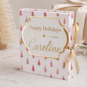 May include: A rectangular white gift box featuring a repeating pattern of small pink Christmas trees. The front displays a gold-foiled label with the text Happy Holidays and the name Caroline in script, finished with a gold ribbon tied on the side.