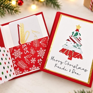 May include: A red and white Christmas greeting card holder featuring a gold foil tree illustration and the text Merry Christmas Kandi & Dan. The interior pocket displays a red pattern with white snowflakes and the words Fa La La.