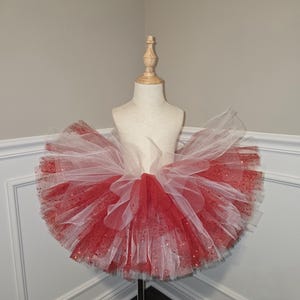 May include: A red and white tutu skirt with glitter accents. The skirt is displayed on a mannequin and is made of layers of tulle. The red sections have glitter, adding sparkle. This tutu is suitable for dance, costumes, or dress-up.