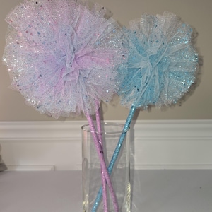 May include: Two decorative wands with glittery tulle pom-poms in pastel colors, one lavender and the other blue, are displayed in a clear glass vase. The wands have matching glitter-covered sticks. The background is a neutral wall.