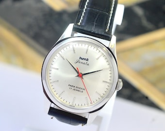 Peut inclure: Montre-bracelet argentée avec un cadran blanc, des aiguilles noires et une trotteuse rouge. Le texte "hmt Janata" et "PARA SHOCK 17 JEWELS" est visible. Bracelet en cuir noir.