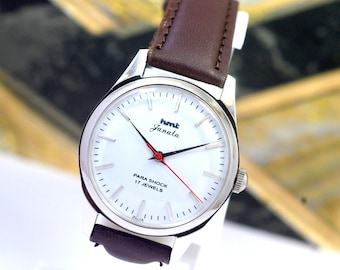 Peut inclure: Une montre argentée avec un cadran blanc et un bracelet en cuir marron. Le cadran affiche le texte "hmt Janata" et "PARA SHOCK 17 JEWELS". L'aiguille des secondes est rouge.